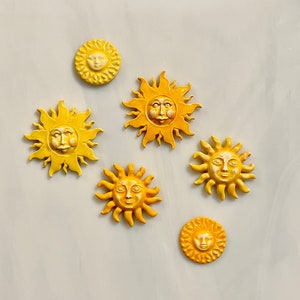 May include: A collection of six yellow and orange sun-shaped decorative items. Each sun has a smiling face and radiating rays. The suns vary in size and shade, creating a cheerful and whimsical display. These decorative items are perfect for adding a touch of sunshine to any space.