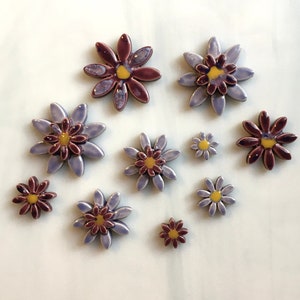 May include: A collection of ceramic daisy flowers in various sizes. The flowers have purple and burgundy petals with yellow centers. The glossy finish gives them a handcrafted look. These decorative flowers are perfect for home decor or craft projects.