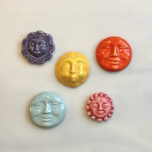 May include: A collection of five ceramic sun and moon face tiles in various colors. The tiles include a purple flower-shaped sun, a yellow face, an orange face, a light blue face, and a pink sun with a scalloped edge.