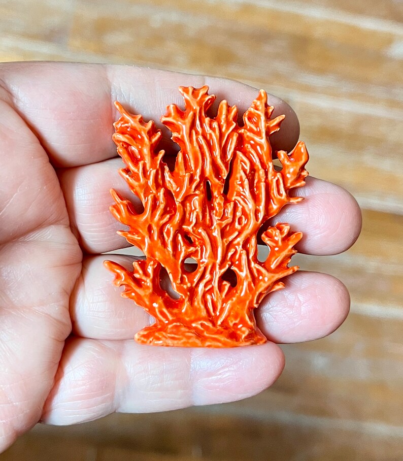 May include: A vibrant orange coral sculpture with intricate details. The coral is a bright, glossy orange, and the design mimics the natural structure of coral reefs. The sculpture is held in a hand, showcasing its size and form.