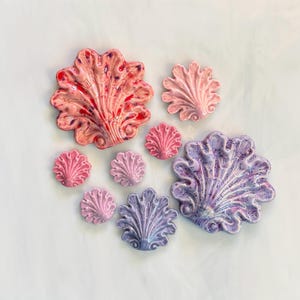 May include: A collection of nine ceramic shell-shaped decorative pieces in varying sizes. The items feature glossy finishes in shades of pink, purple, and lavender, with some pieces displaying speckled or marbled color patterns across their surfaces.