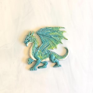 May include: A decorative dragon figurine featuring a glossy finish in shades of turquoise, teal, and lime green. The creature is depicted in a standing pose with large, detailed wings spread wide, a long curved tail, and a textured, scaled body.