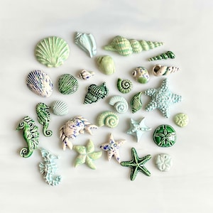 May include: A collection of ceramic sea life figurines, including seashells, starfish, seahorses, and a crab. The figurines are in shades of green, blue, and white, with intricate details and textures. The arrangement is on a white surface.