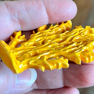 May include: A bright yellow, coral-shaped decorative object. The object has a textured surface with a glossy finish. The coral-like design is held in a person's hand, with a blurred wooden background.