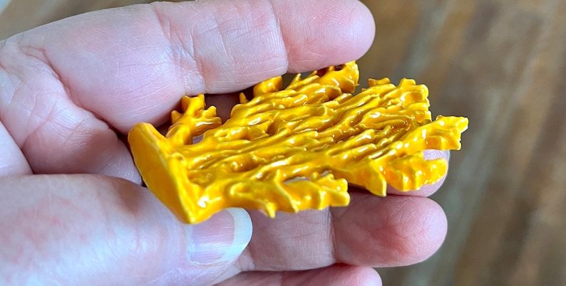 May include: A bright yellow, coral-shaped decorative object. The object has a textured surface with a glossy finish. The coral-like design is held in a person's hand, with a blurred wooden background.