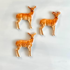 May include: Three ceramic deer figurines in shades of brown and white are arranged against a light blue background. The deer have detailed features, including antlers and spotted patterns on one figurine. These decorative pieces are ideal for home decor.