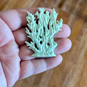 May include: A light green ceramic coral sculpture with intricate details. The coral has a textured surface and a base for stability. The sculpture is held in a hand, showcasing its size and form. The color is a soft, muted green.