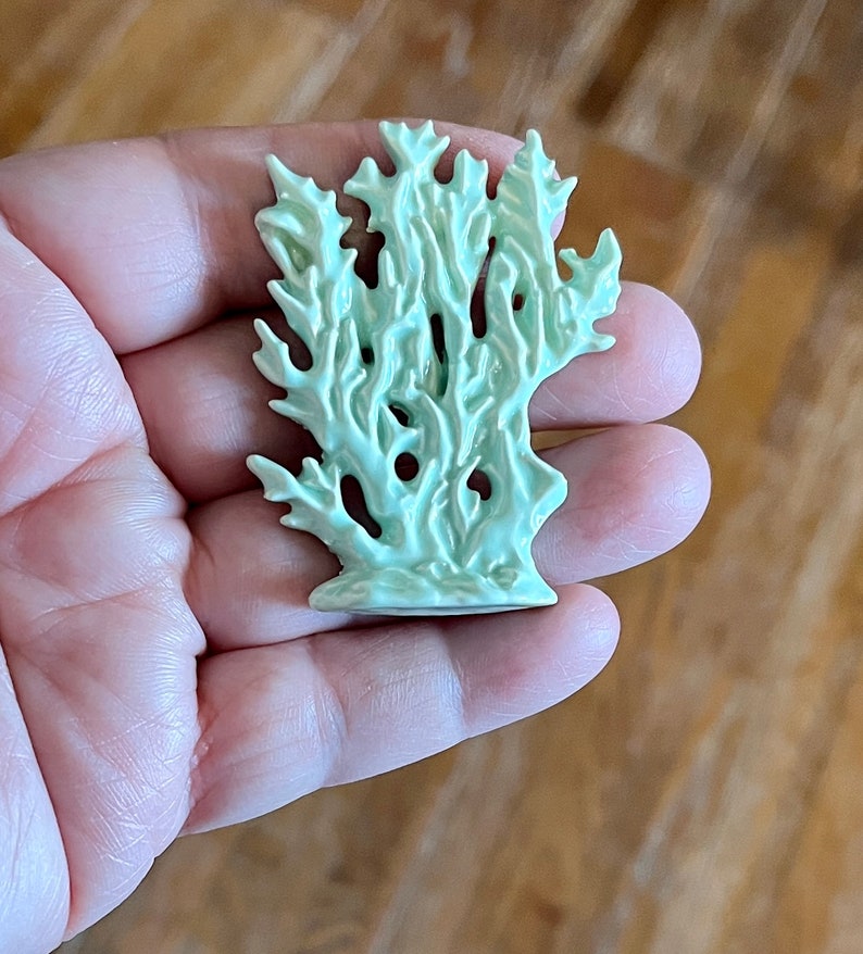 May include: A light green ceramic coral sculpture with intricate details. The coral has a textured surface and a base for stability. The sculpture is held in a hand, showcasing its size and form. The color is a soft, muted green.