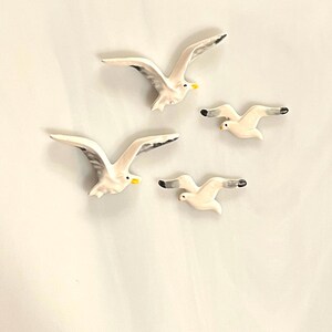 May include: Four ceramic seagull figurines in flight. The seagulls are white with gray wing tips and yellow beaks. Two are larger, with outstretched wings, and two are smaller, with wings partially folded.