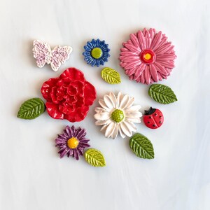 May include: A collection of ceramic decorative pieces including a white butterfly, a small blue flower, a large pink dahlia, a red rose, a white daisy, a purple aster, a red ladybug with black spots, and four green leaves with textured veins.