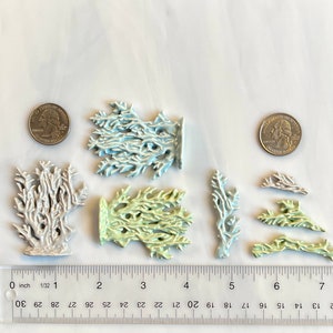 May include: A collection of ceramic coral sculptures in various sizes and colors, including white, light blue, and pale green. A ruler shows measurements in inches, and two quarters are included for scale. The coral pieces have intricate, branch-like designs.