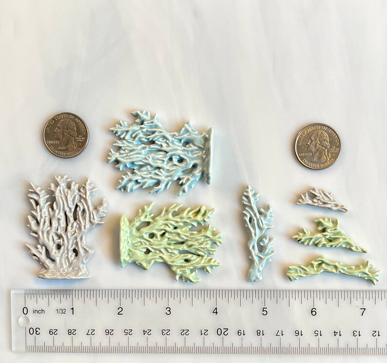 May include: A collection of ceramic coral sculptures in various sizes and colors, including white, light blue, and pale green. A ruler shows measurements in inches, and two quarters are included for scale. The coral pieces have intricate, branch-like designs.