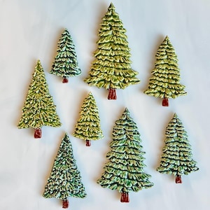 May include: A collection of nine ceramic Christmas tree ornaments. The trees are various sizes, painted in shades of green with white accents, and have red bases. The ornaments are arranged on a white background.