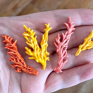 May include: Four colorful coral-shaped decorative pieces in shades of orange, yellow, and pink. The pieces have a detailed, textured surface and are arranged in a hand. These decorative pieces are likely for crafts or home decor.