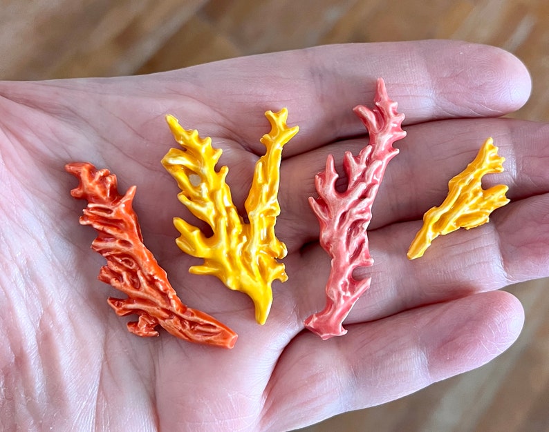May include: Four colorful coral-shaped decorative pieces in shades of orange, yellow, and pink. The pieces have a detailed, textured surface and are arranged in a hand. These decorative pieces are likely for crafts or home decor.
