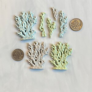 May include: A collection of ceramic coral sculptures in various sizes and colors, including light blue, green, and silver. The coral pieces are arranged on a white surface, with two quarters for size reference. The sculptures have a glossy finish.