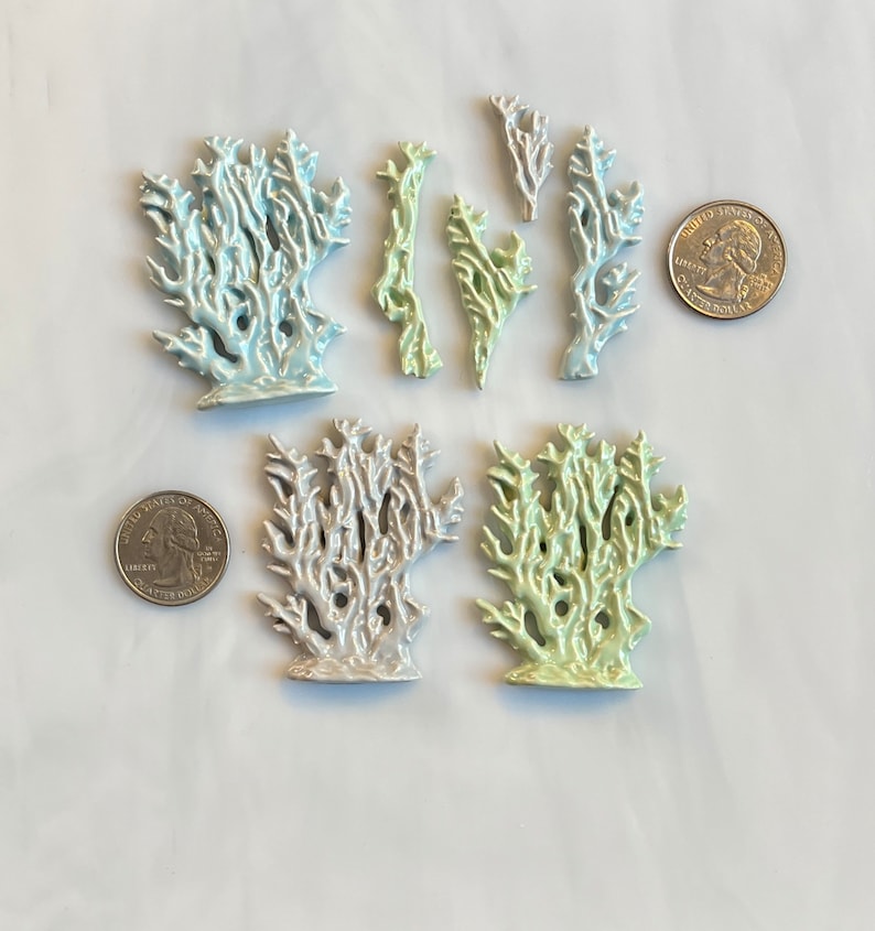 May include: A collection of ceramic coral sculptures in various sizes and colors, including light blue, green, and silver. The coral pieces are arranged on a white surface, with two quarters for size reference. The sculptures have a glossy finish.