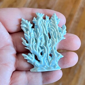 May include: A light blue ceramic coral sculpture with intricate details. The coral has a textured surface and a glossy finish. The sculpture is held in a hand, showcasing its size and form. The piece is likely a decorative item.