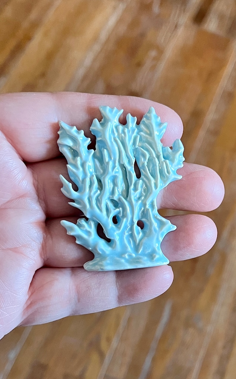 May include: A light blue ceramic coral sculpture with intricate details. The coral has a textured surface and a glossy finish. The sculpture is held in a hand, showcasing its size and form. The piece is likely a decorative item.