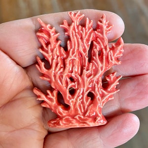 May include: A coral-colored ceramic sculpture of a coral reef. The sculpture has a detailed, textured surface with many branches and holes. The coral is a vibrant shade of orange-red, and the overall design is reminiscent of marine life.