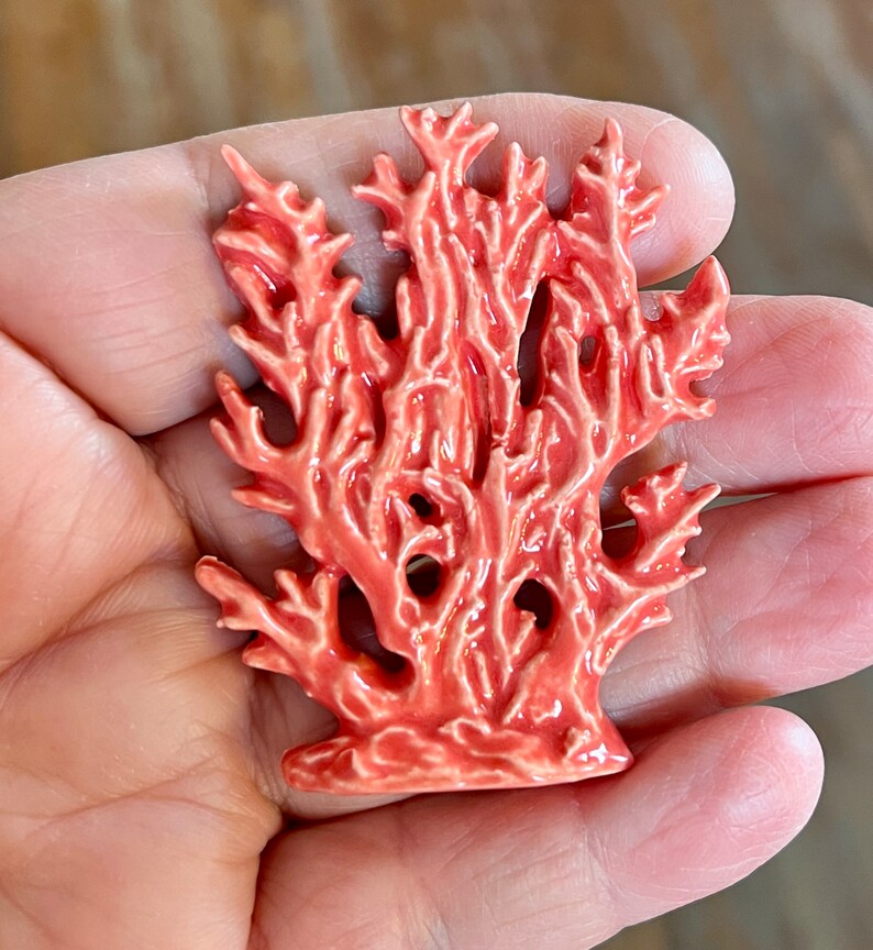 May include: A coral-colored ceramic sculpture of a coral reef. The sculpture has a detailed, textured surface with many branches and holes. The coral is a vibrant shade of orange-red, and the overall design is reminiscent of marine life.