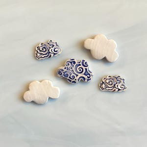 May include: Five cloud-shaped ceramic ornaments. Two are white, and three are white with blue swirling patterns. The ornaments are arranged on a light blue surface. These decorative pieces could be used for home decor or craft projects.