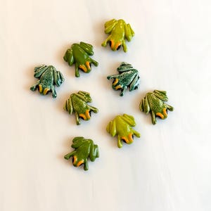 May include: Eight small frog-shaped buttons in various shades of green. Each frog features a smooth, glossy finish with distinct orange accents around the eyes and black pupils. The frogs are arranged in a scattered pattern, showing different textures.