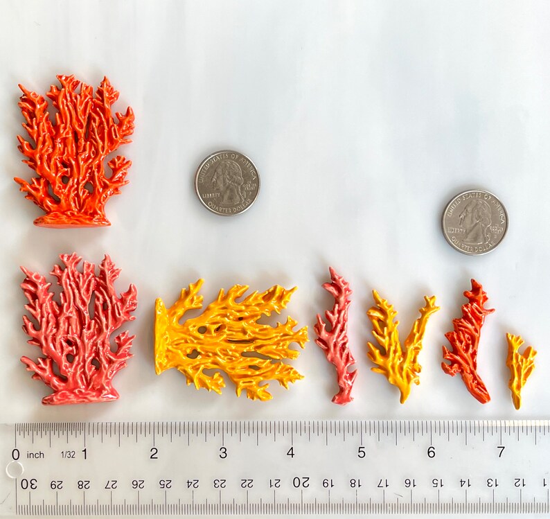 May include: A collection of colorful ceramic coral sculptures in shades of orange, red, and yellow. The coral pieces vary in size and shape, with a ruler showing measurements in inches. Two quarters are placed near the coral for size comparison.
