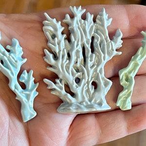 May include: Three ceramic coral sculptures in shades of blue, white, and green. The coral pieces are intricately detailed, with a textured surface and a glossy finish. The sculptures are displayed on a hand.