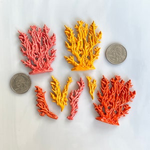 May include: A collection of coral-shaped ceramic ornaments in shades of orange, yellow, and pink. The ornaments vary in size and shape, with intricate branch-like details. Two quarters are included for size reference. The coral is arranged on a white surface.