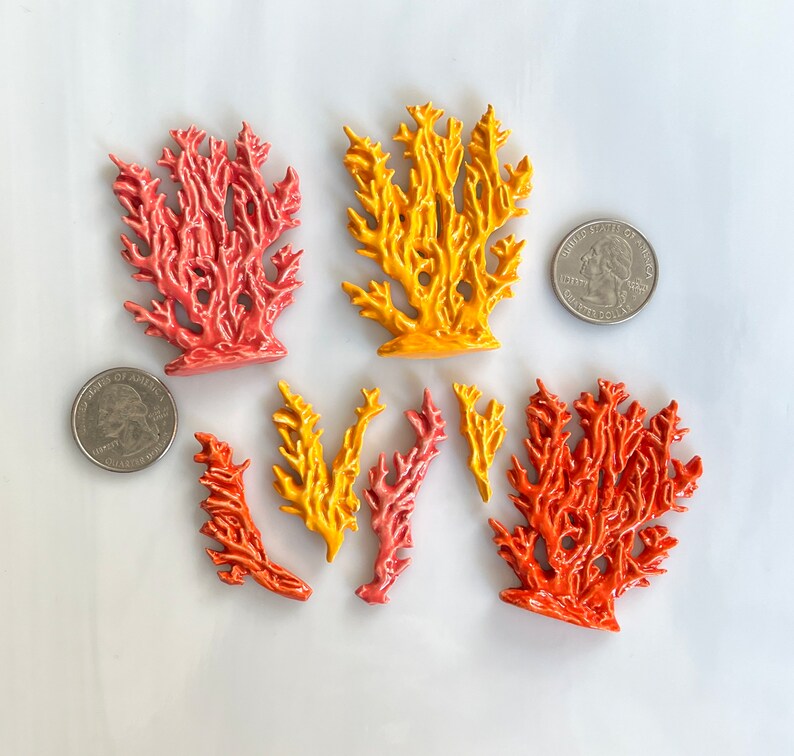 May include: A collection of coral-shaped ceramic ornaments in shades of orange, yellow, and pink. The ornaments vary in size and shape, with intricate branch-like details. Two quarters are included for size reference. The coral is arranged on a white surface.