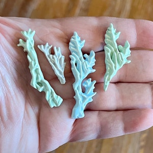 May include: Four small, decorative coral-shaped pieces in shades of pale green, blue, and white. The ceramic pieces have a textured surface and are arranged in a hand.