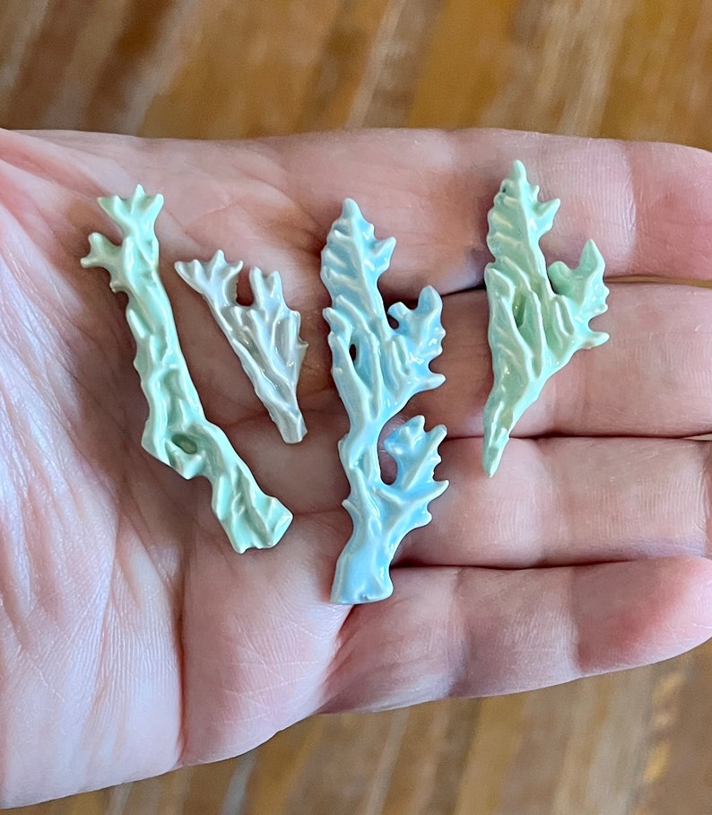 May include: Four small, decorative coral-shaped pieces in shades of pale green, blue, and white. The ceramic pieces have a textured surface and are arranged in a hand.
