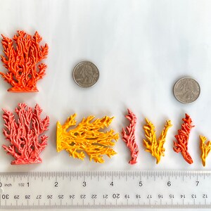 May include: A collection of colorful ceramic coral sculptures in shades of orange, yellow, and pink. The coral pieces vary in size and shape, with a ruler showing measurements in inches. Two quarters are placed near the coral for size comparison.