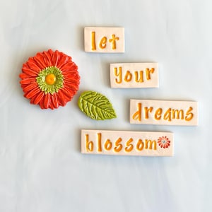 May include: A collection of ceramic decorative pieces, including a vibrant red and yellow flower, a green leaf, and rectangular tiles with the words "let your dreams blossom" in orange lettering. The pieces are arranged on a light blue surface.