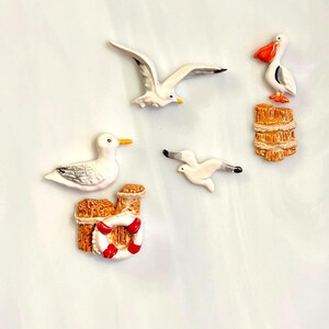 May include: A set of five nautical-themed magnets featuring two white seagulls with gray wings, one flying and one perched, a small flying white gull, a pelican perched on a wooden piling, and a wooden piling with a red and white life ring.