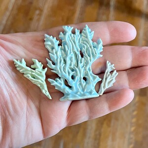 May include: Three ceramic coral sculptures in shades of blue and green are displayed in a hand. The largest piece is a light blue coral formation, with two smaller green and white coral pieces. The sculptures have a textured surface.