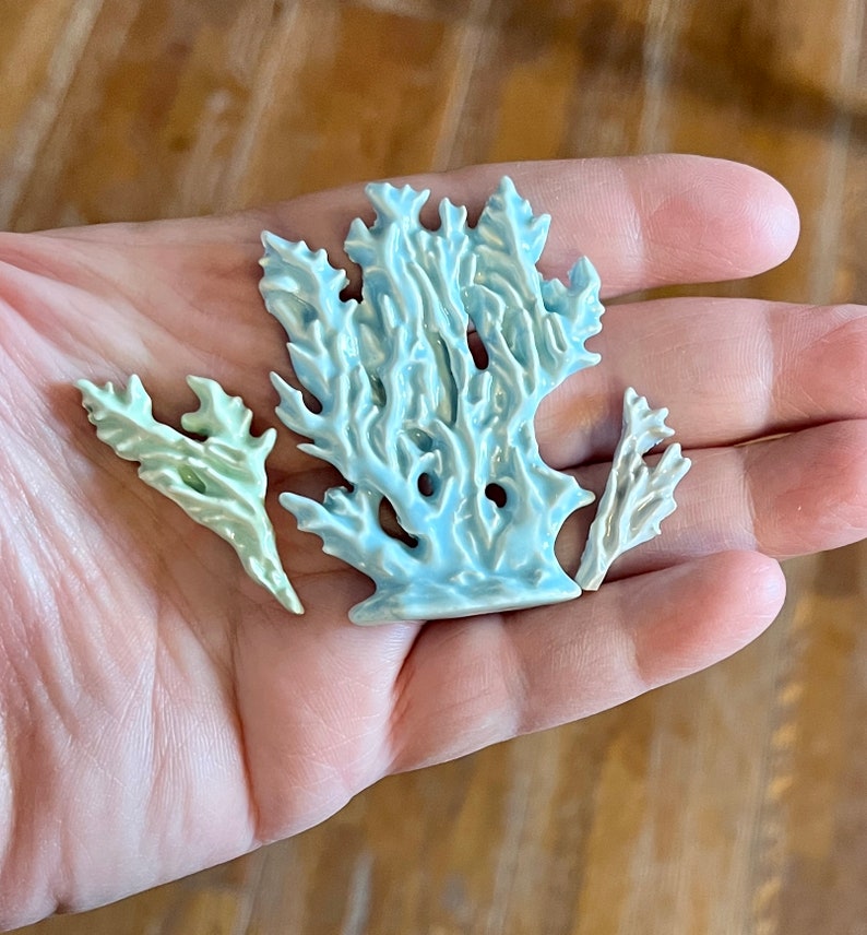 May include: Three ceramic coral sculptures in shades of blue and green are displayed in a hand. The largest piece is a light blue coral formation, with two smaller green and white coral pieces. The sculptures have a textured surface.