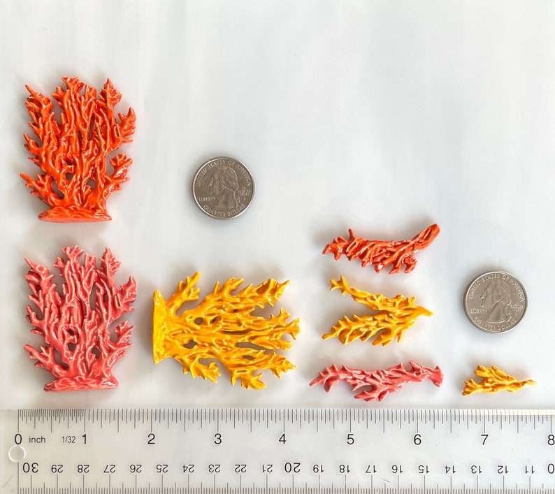 May include: A collection of coral-shaped ceramic pieces in various sizes and colors, including orange, yellow, and pink. A ruler and two quarters are included for size reference. The coral pieces are arranged on a white surface.