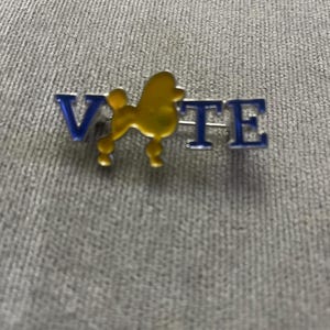 May include: A pin with the word "VOTE" in blue letters. The "O" is replaced with a yellow poodle silhouette. The pin is made of metal and has a shiny finish. The pin is on a gray fabric background.