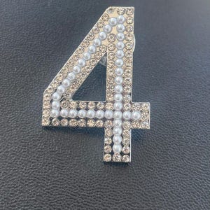 May include: A silver-toned number four brooch decorated with white pearls and rhinestones.