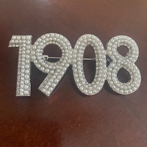 May include: A silver-toned brooch shaped like the number 1908, decorated with white pearl-like beads.