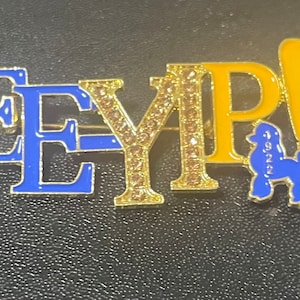 Könnte beinhalten: Goldfarbene Brosche mit blauem Emaille und Strasssteinen, die "EEYP!" buchstabieren, mit einer kleinen blauen Pudelfigur neben dem Ausrufezeichen.