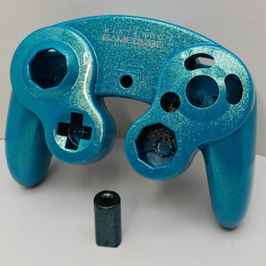 May include: A turquoise glitter-finish Nintendo GameCube controller shell with empty button slots and a matching small cylindrical component. The text NINTENDO GAMECUBE is embossed on the front surface of the controller housing.