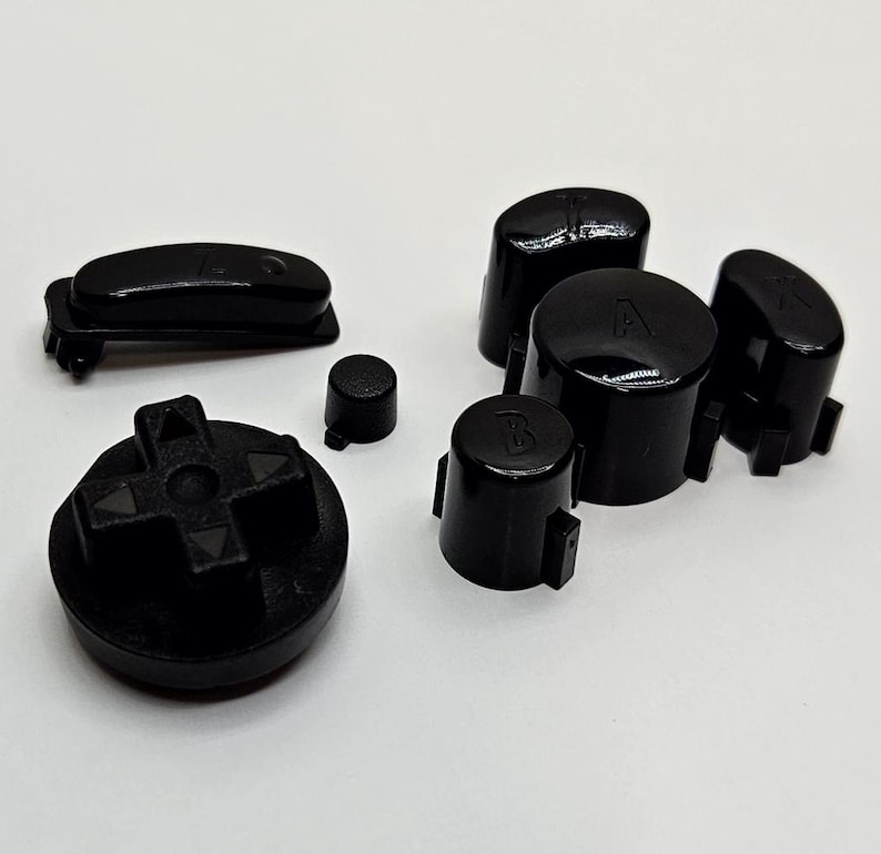 May include: A set of black replacement buttons for a video game controller. The set includes a directional pad, four face buttons, a start button, and a select button.