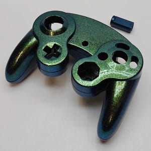 May include: A metallic color-shifting game controller front shell with a green and blue iridescent finish, featuring cutouts for buttons and joysticks, accompanied by a small, matching rectangular component piece.
