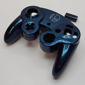 May include: A dark blue metallic gaming controller shell with a glittery finish. The front face features the Super Smash Bros logo engraved in white. The shell includes cutouts for buttons, joysticks, and a small separate rectangular piece.