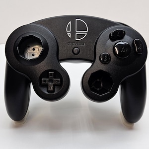 May include: A black Nintendo GameCube controller with a white Nintendo logo and the words "START/PAUSE" printed on the top. The controller has a black D-pad, two black analog sticks, and four black buttons.