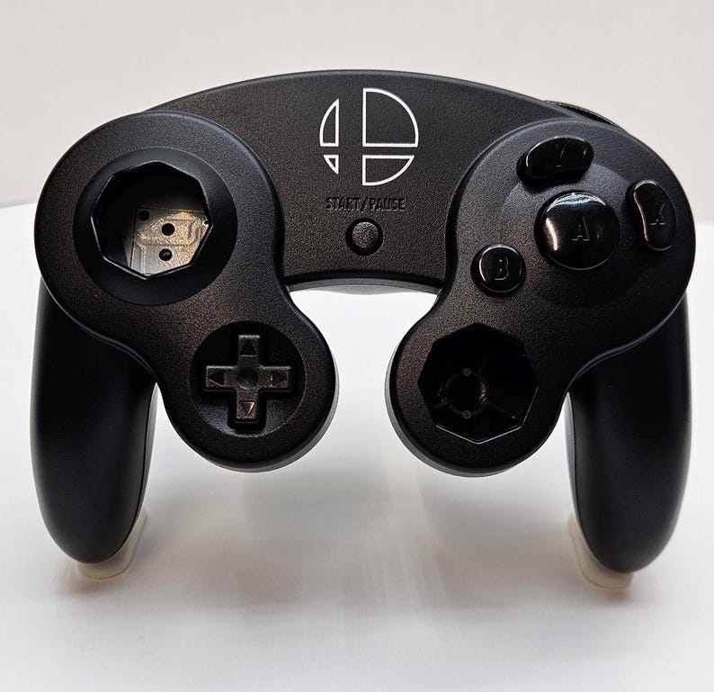 May include: A black Nintendo GameCube controller with a white Nintendo logo and the words "START/PAUSE" printed on the top. The controller has a black D-pad, two black analog sticks, and four black buttons.