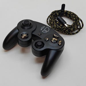 Puede incluir: Mando de videojuegos negro con detalles dorados en los botones y un cable negro y dorado a juego. El mando tiene un diseño único con un logotipo. El cable está enrollado y tiene un conector USB.